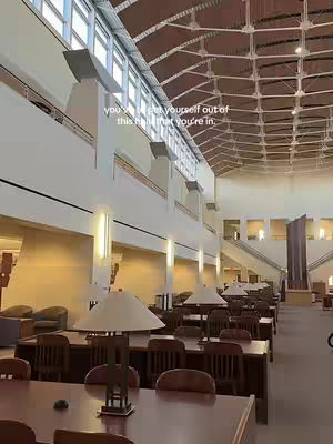 A wide shot of a library interior. The space features a high ceiling with a complex network of beams and skylights. Rows of tables with lamps and chairs are arranged throughout the room. The walls are a neutral color, and the natural light from the windows and skylights illuminates the space.