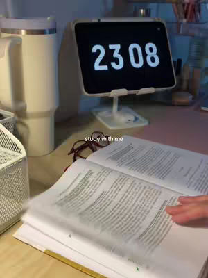 An overhead shot of a desk setup with a book open, an electronic clock displaying the time 23:08,  a large white tumbler, a wire basket, and a pair of eyeglasses. The book's pages are being turned by a hand out of the frame. The setting suggests a study environment.