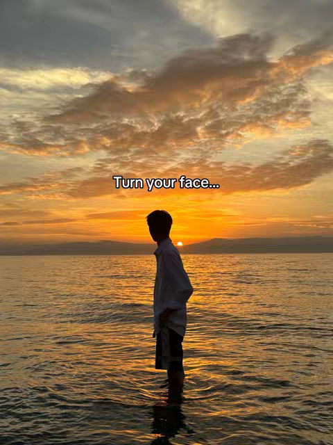A silhouetted figure of a person standing in the water at sunset. The sky is filled with orange and yellow hues from the setting sun, with clouds scattered about. The person is standing in the water, facing the sunset. Text overlay reads: "Turn your face..."