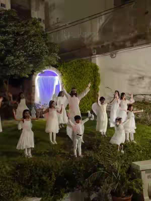 The image shows a group of people, likely children and adults, in a garden setting during what seems like an evening or night. They are all dressed in white or light-colored robes. A central figure, possibly portraying a religious figure like Jesus, stands with arms outstretched. Surrounding figures have their arms raised as well. There's a backdrop suggesting a structure with a light emanating from within. Overall, the image depicts a religious or theatrical performance, with a focus on the central figure, and everyone else in the background.
