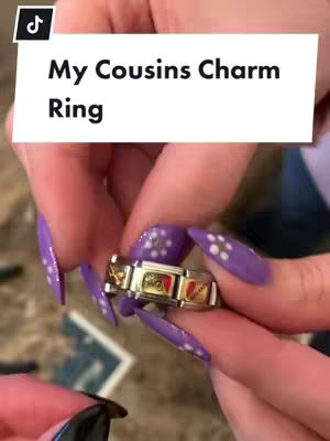 A close-up shot of a hand with long, pointed, purple nails holding a gold and jewel-adorned charm bracelet.