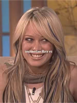 A medium shot featuring a woman with long blonde hair, smiling, and engaging the viewer. The text "september libra vs" is overlayed on the image. The background appears to be a blurred interior setting, possibly a studio or a set.
