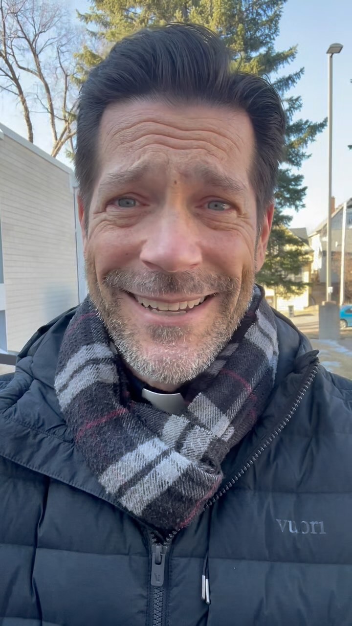 A close-up shot features a smiling man with short, dark hair and a trimmed beard. He's wearing a black jacket and a patterned scarf. The background is blurred, indicating an outdoor setting with a building visible on the left.
