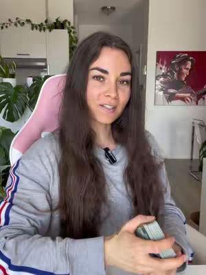 A woman with long brown hair smiles while holding a deck of cards. She is wearing a gray sweater, and there is a microphone clipped to her shirt. Behind her is a white wall, plants, and a painting on the wall.