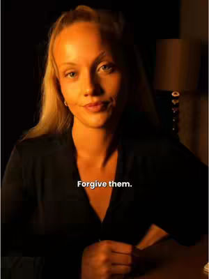 A woman with blond hair and a black top is centered in a slightly blurry, warm-toned image. The lighting is soft, appearing to come from the side, highlighting her face and shoulders. She is looking at the camera with a slight, knowing smile. Below her, the overlaid text 'Forgive them.' is visible.