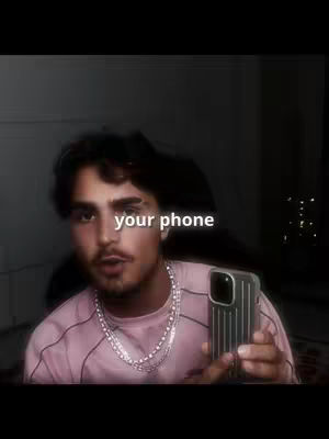 A man is holding a phone while looking directly into the camera. The image is medium shot, with the foreground featuring the man's face, and the background is dark. Text overlayed across the image reads 'your phone'. The man wears a pink shirt and a silver necklace