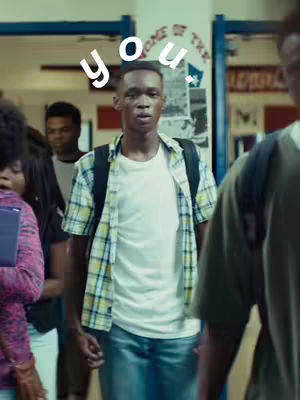 A middle-aged Black man wearing a plaid shirt over a white t-shirt, jeans, and a black backpack, standing in a hallway with other students partially visible in the background. The words 'you.' are superimposed over the man's head in white.