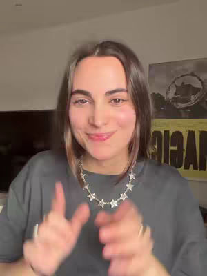 A woman is in a selfie-style video, smiling at the camera. Her hands are raised, palms facing out and slightly angled. She is wearing a gray t-shirt and a necklace with star-shaped charms. A poster is visible in the background.