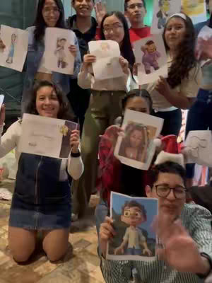 A group of people, seemingly at an event, hold up drawings of various cartoon characters and sketches. The people are of diverse ages and genders, dressed in casual attire. The background is slightly blurred, suggesting an indoor setting, with hints of decorations.