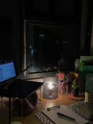 A nighttime study scene with a laptop, candle, and notebook on a desk. The window shows city lights in the background.
