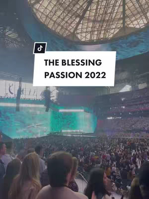 A wide shot of a large event in a stadium. A huge crowd of people are gathered, likely at a large concert or event. The lighting is bright, focused on a large screen in the background, and creating an overall sense of excitement. A large rectangular white banner with black text is placed on the screen with the text "THE BLESSING PASSION 2022".