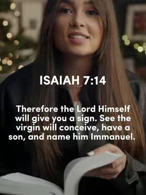 A woman with long brown hair is holding an open book. The text overlay shows Isaiah 7:14, with the verse about the virgin conceiving a son. The background is softly blurred with visible Christmas lights.
