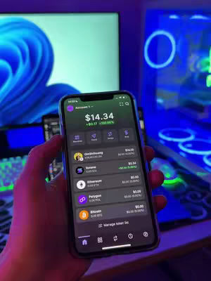 A person holding a smartphone displaying a trading app interface in front of a computer monitor and a computer tower with neon lights. The person's hand and the phone are in the foreground, with the screen of the phone showing account information.