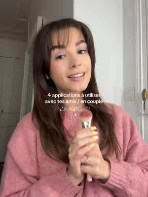 A woman with long brown hair and bangs is centered in the frame, holding a makeup brush. She wears a pink sweater and has rings on her fingers. Text overlayed reads, "4 applications à utiliser avec tes amis / en couple". The background is a plain white wall with a door visible.