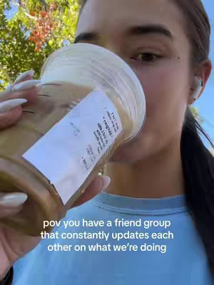 A selfie-style shot featuring a woman holding a Starbucks coffee. She is seen from the chest up, with the coffee cup obscuring a portion of her face. The background shows a blurry mix of green foliage and sunlight. The woman, wearing earbuds, seems to be in an outdoor setting, with the focus primarily on her and the coffee.