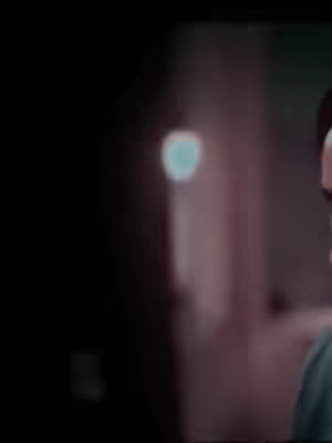 The image is slightly blurred, with a dark background that contrasts with a lighter, possibly indoor setting. There is a blurred figure visible, and the overall color palette appears to be muted, with soft hues. The light source seems to be coming from front, creating a sense of depth and atmosphere.
