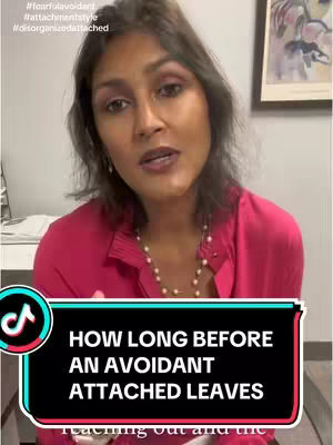 A woman with dark hair and light skin is centered in the frame, wearing a bright pink button-down shirt and a pearl necklace. She speaks toward the camera. A text overlay at the bottom of the screen has the text: "HOW LONG BEFORE AN AVOIDANT ATTACHED LEAVES". Behind her, there is a wall and what looks like a framed picture. The background is blurred, focusing attention on the subject and the overlay.
