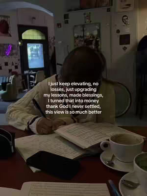 A person is seated at a table, hunched over with a book and papers, pen in hand, presumably writing or taking notes. A coffee cup, phone, and writing supplies also are visible and in the background is a doorway and a portion of a window offering a view of a building's interior and exterior.