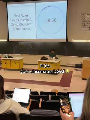 A classroom setting with a professor standing in front of a projector screen. The screen displays rules for a final exam, and a timer. Students are seated in rows, with some using laptops and phones. The overall impression is casual and hints at a relaxed academic atmosphere.