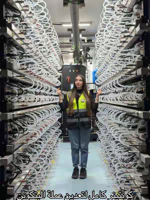 A full-shot image in an industrial setting. A woman stands between a large white structured machine. The machine is composed of many lines and tubes. The woman is wearing a yellow vest over a black shirt and blue jeans. The lighting is somewhat dim with artificial lights overhead. Text in Arabic script is on the bottom of the image.