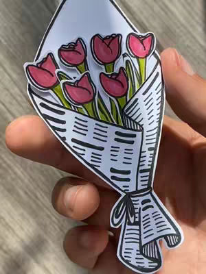 A close-up shot of a paper bouquet of tulips held in a person's hand. The bouquet is made out of paper and has a white background. The tulips are pink and green. The bouquet is wrapped in newspaper.