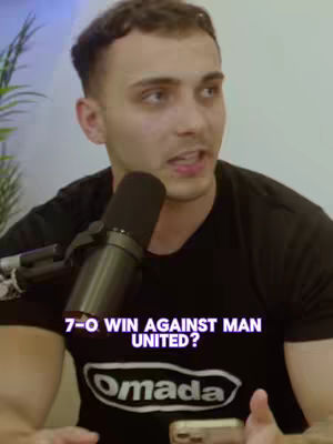 A man is speaking into a microphone in front of a light background. The text overlay reads "7-0 WIN AGAINST MAN UNITED?" in a white font. He wears a black t-shirt with the brand "Omada" displayed prominently on the front. His hair is short and styled, he is fair-skinned, and has a focused expression. A smartphone is in his hand.