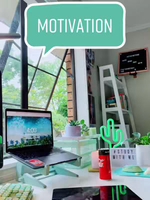 The image shows a well-lit desk with a laptop, study supplies, and decorative items. A laptop on a stand is the focal point, displaying a screen with blue and white graphics. A speech bubble with the word "MOTIVATION" is overlaid. Various other items, including a can of Coca-Cola, a cactus-shaped neon light, and a sign that reads "#STUDY WITH ME", are visible, giving a sense of a study-focused environment. The overall composition is clean and inviting.