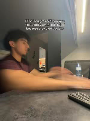 A man is sitting at a desk in front of a blurry background, seemingly studying. On the desk is a black keyboard and a bottle of water.