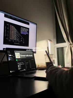 A dimly lit scene of a study setup with a laptop displaying an app related to music while a large screen displays a graph. The scene is captured from a low angle, showing a partially visible hand holding a pen near a cup of coffee. The overall composition is minimalist, with muted tones and a focus on the digital interface.