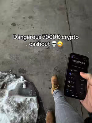 A person's feet are shown standing on a gray, textured surface, with snow in the lower left corner. The individual is wearing brown boots and gray jeans. A smartphone is held in the person's right hand displaying a crypto account balance. Text overlay indicates caution about a crypto cashout.