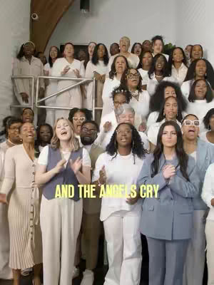 A large group of people, primarily women, dressed in light-colored clothing, sing in what appears to be a church or auditorium. They are positioned on multiple levels, with some standing behind and others in the foreground. The text overlay says "AND THE ANGELS CRY". The overall scene suggests a choir performance or a worship service.