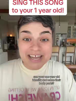 A selfie-style video cover shot featuring a woman with short, dark hair. Above her head is a white rectangular text box with red text, "SING THIS SONG to your 1 year old!". Below her, there's another text on a semi-transparent rectangular box that reads "can your one year old identify and name their body parts?". The woman is smiling with her mouth open, and the background shows a living room.