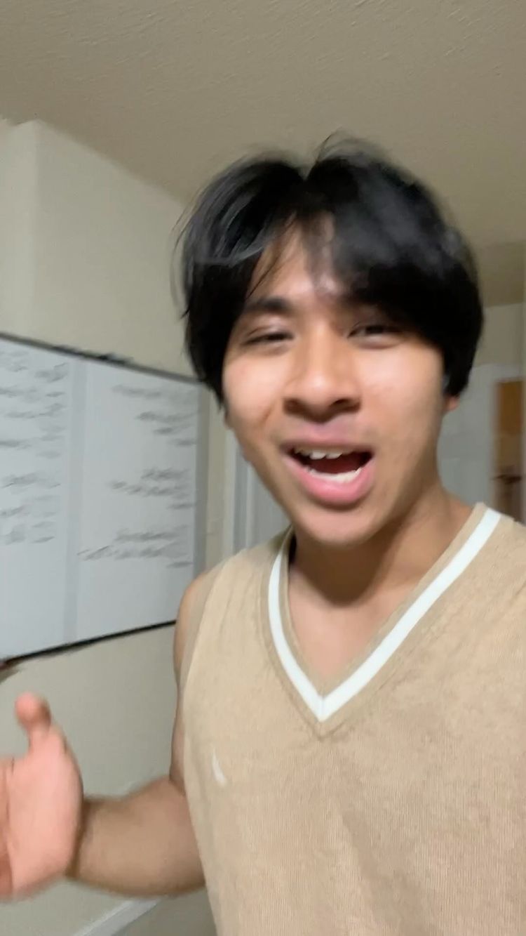 A young man with dark hair and a tan vest smiles and gestures in a room. A whiteboard with writing is partially visible behind him.