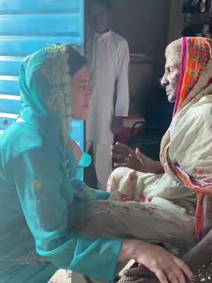 The image shows two women in a close-up conversation. The woman on the left is wearing a turquoise headscarf and a matching top, her face angled towards the other woman. The woman on the right is older, with a patterned headscarf and a white outfit, and is seated. They appear to be in an interior setting, possibly a home, with a man in a white shirt in the background.