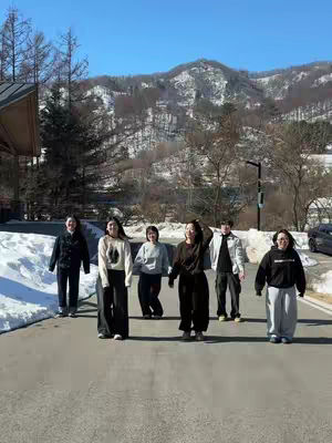 A group of six people, likely friends, are walking towards the camera on an asphalt road. Behind them are snowy mountains and a clear blue sky. They appear to be dressed for cold weather. The lighting suggests it's a bright, sunny day.