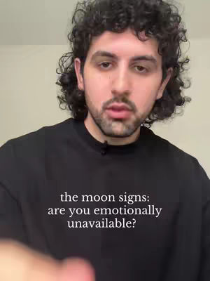 A man with curly dark hair and a beard is centered in the frame, wearing a black sweatshirt. The background is a plain, off-white wall. Text overlaid on the sweatshirt reads, "the moon signs: are you emotionally unavailable?"
