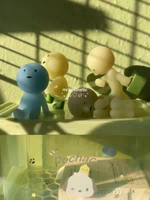The image features various small, abstract figures arranged in a whimsical scene. A blue figure with a round head sits towards the left. Adjacent to it, there are two yellow, vaguely mushroom-shaped figures, and a rounded, grey figure. To the right, a taller figure poses with outstretched arms. The figures are set against a soft, gradient background with what appears to be a window. Underneath this upper layer, a cuter, cartoon-like image of a white character with a small hat is present.
