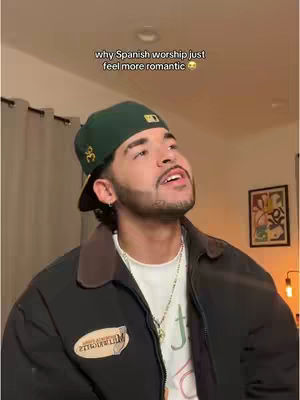 A man with a beard and a green cap is looking up with a light smile, set against a room with a painting and warm lighting.
