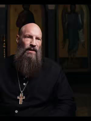 A bald man with a thick, gray beard wearing a black shirt and a cross necklace. He looks slightly to the side with a serious expression. Behind him are blurred paintings.