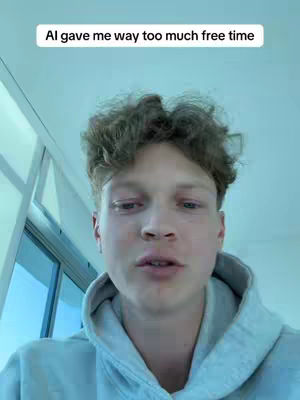 A young man with curly hair, wearing a grey hoodie, is centered in the frame, looking directly at the camera. The background is a light blue, suggesting an indoor setting. Above the man's head, a white banner with black text reads 'Ai gave me way too much free time'. The overall composition gives a sense of casual filming.