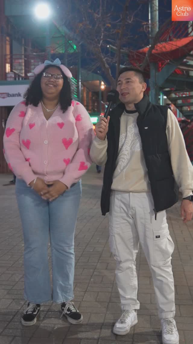 Two people are standing outdoors for an interview. The person on the left is wearing a pink sweater with heart details and a headband, and the person on the right is holding a microphone.