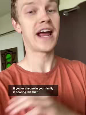 A man with light skin and light brown hair is speaking while looking at the camera. The background suggests an interior setting with a partially visible door and a framed artwork. A text overlay at the bottom states, "If you or anyone in your family is snoring like that." The man is wearing an orange t-shirt, and his hand is visible in the foreground.