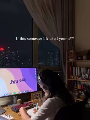 A nighttime interior shot featuring a person working at a desk, with a cityscape visible through a window. A text overlay at the top reads 'If this semester's kicked your a**'. The person wears headphones and is focused on studying, with a computer monitor displaying text that reads 'you can'.