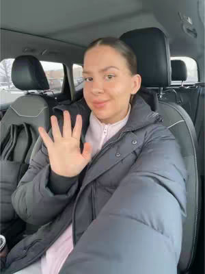 A person sitting in the front seat of a car, waving at the camera with their right hand. They are wearing a black jacket and a light pink shirt. The background shows the interior of the car and what looks like a snowy outside scene