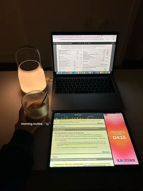 A top-down shot of a desk setup featuring a laptop, tablet, a cup of coffee, and a lit lamp. The laptop displays a document, while the tablet shows a screen with notes and a timer. The overall aesthetic is minimalistic, with a warm lighting.
