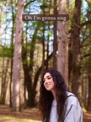 A woman with long dark hair smiles in a forest, with tall trees in the background. The image has a warm, earthy tone.