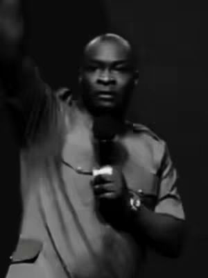 A grayscale image of a man in a button-down shirt holding a microphone with his right hand and raising his left arm. The background is dark and blurred, focusing the viewer's attention on the man's face and gestures.