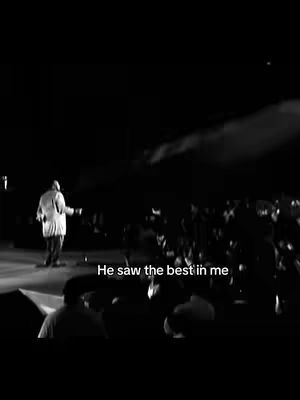 A black-and-white photo depicts a person on a lighted stage addressing a large crowd. The person is seen from behind, wearing a long coat. Below the person on the stage, a caption in white text that reads "He saw the best in me". The crowd is visible but blurred, suggesting a focus on the performer.