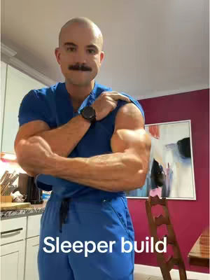 A muscular man with a shaved head and a mustache flexes his arms, wearing a blue scrub top and pants. The background includes a neutral-colored wall, a painting, and a glimpse of a kitchen setup.
