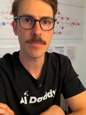 A man with glasses and a mustache looks directly at the camera in a selfie-style shot. He wears a black t-shirt with white text that appears to be a logo. Behind him, a whiteboard or poster with a network diagram is partially visible.
